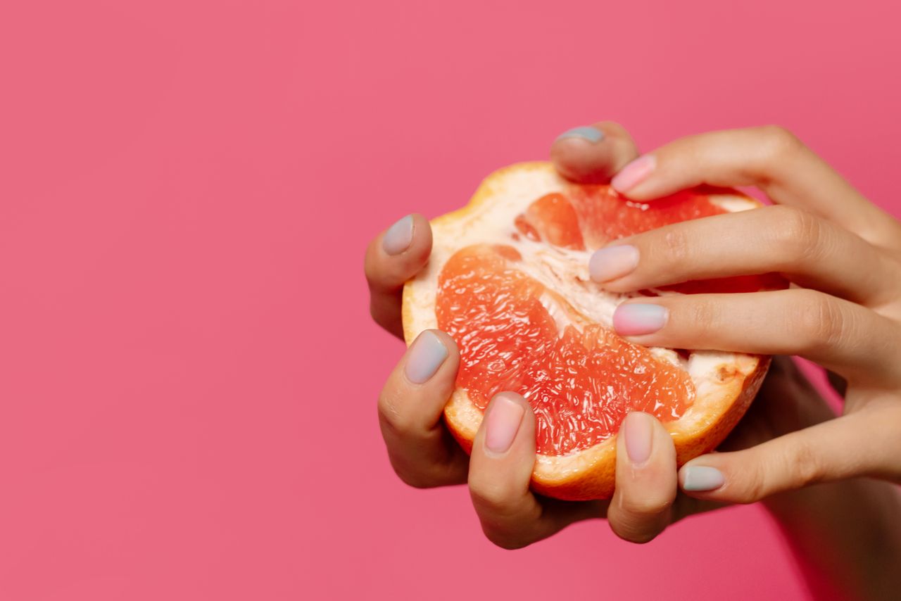 Close-up of hands holding a sliced grapefruit against a pink background, highlighting vibrant colors.